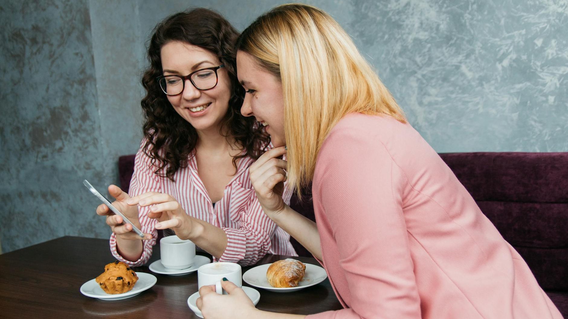 Two women smile over coffee and pastries while one woman shows something on her phone to the other