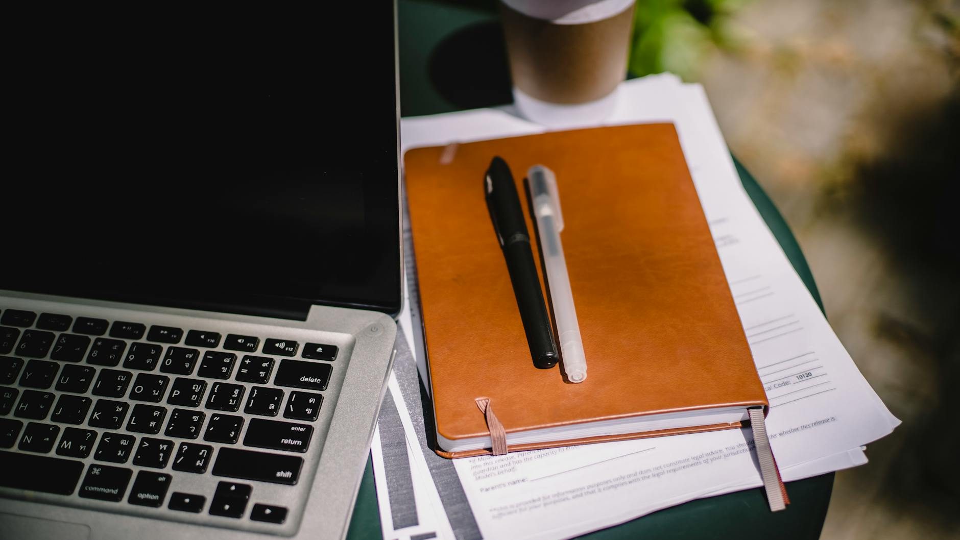 A laptop sits open next to a notebook with pens on top