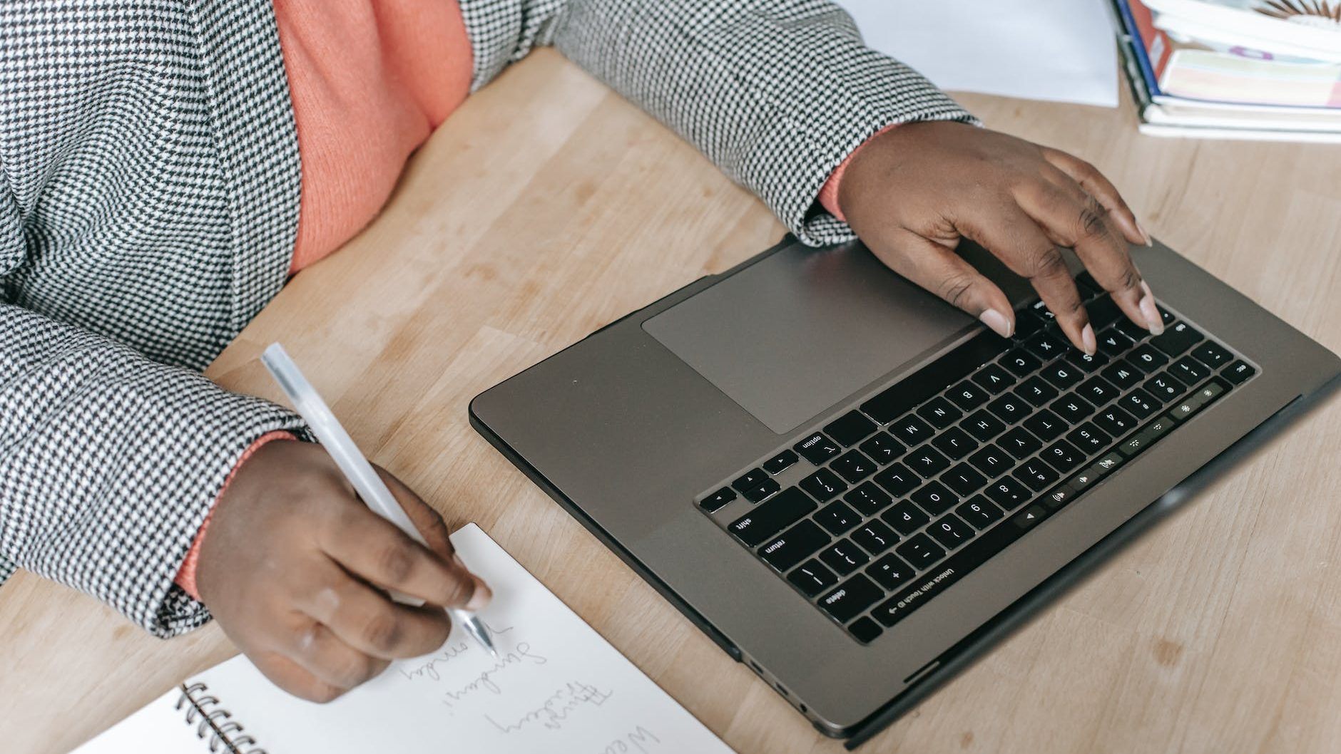 A person types on their laptop with one hand while writing in a notebook with the other